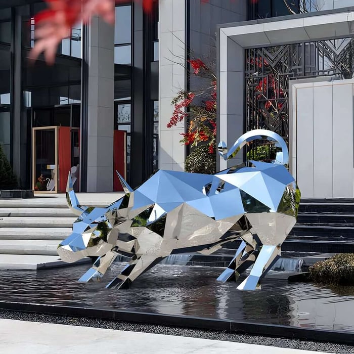A mirrored, faceted metal bull sculpture stands in a shallow reflecting pool in front of a modern building entrance with steps and glass doors.