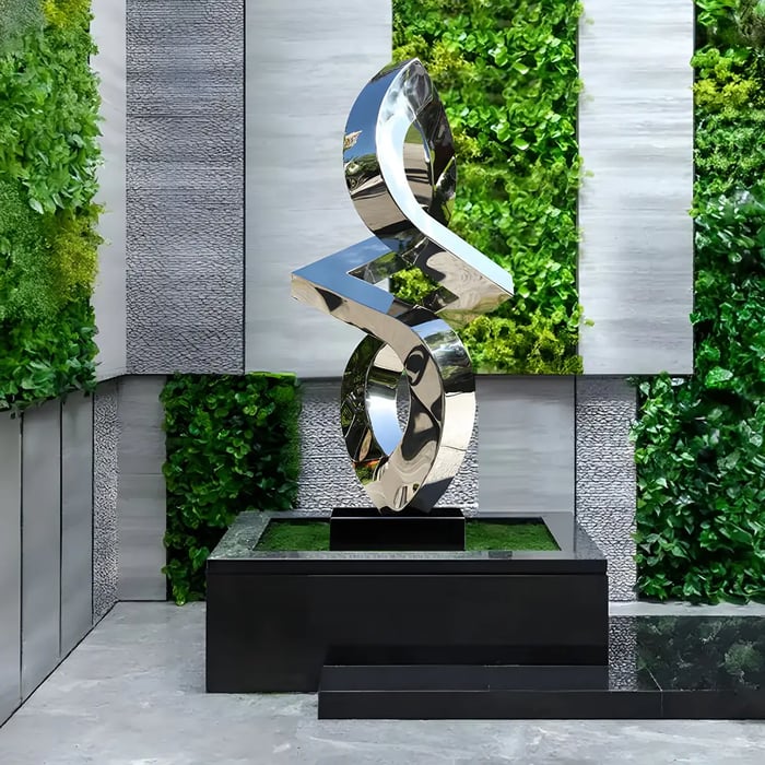 A contemporary abstract stainless steel sculpture with a highly reflective surface, mounted on a black pedestal in a landscaped area with lush greenery.