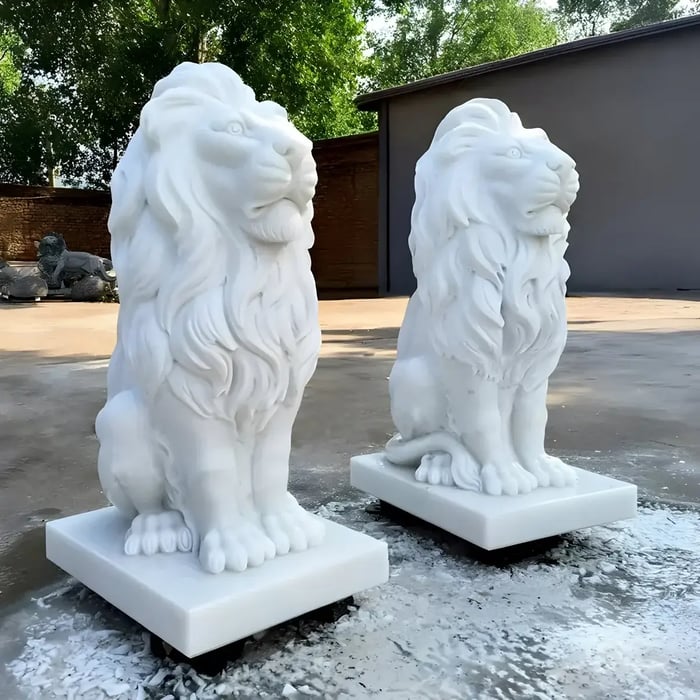 Pair of classic white lion statues seated on square bases, showcasing regal form and symmetry in a sunlit courtyard space.