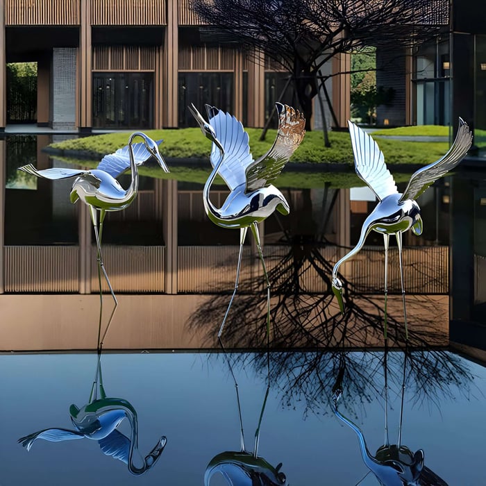 Metallic crane sculptures with spread wings reflected in water, placed in a tranquil courtyard with modern architecture.