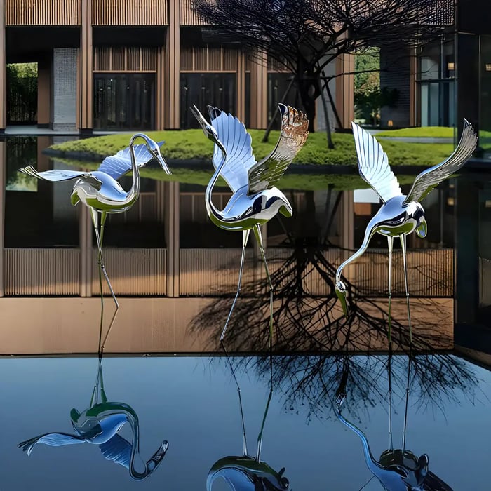  Three polished stainless steel crane garden sculptures with outstretched wings, positioned in a tranquil water feature, reflecting the surrounding architecture and landscape.