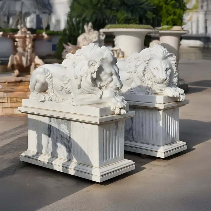 Pair of white lying lion statues on pedestals displayed in an outdoor garden showroom.
