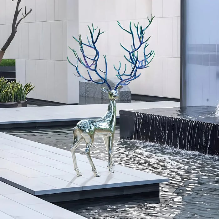 A polished stainless steel deer sculpture with uniquely designed blue antlers, standing beside a modern water feature in a contemporary architectural setting.