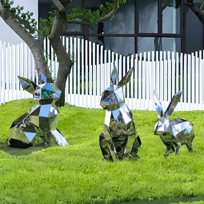 Three faceted mirror stainless steel rabbit sculptures on bright green lawn near a white fence.