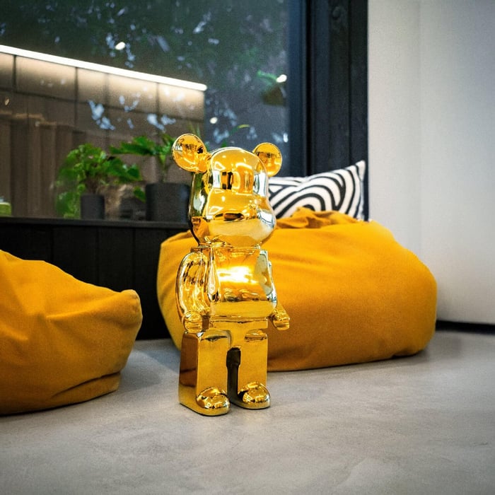 Modern interior with a shiny Gilded Gold Bear Sculpture, 50cm tall, placed between mustard beanbags on a polished grey floor near a window with plants.
