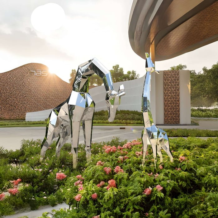Life-size animal sculptures of a giraffe and antelope displayed in a landscaped garden with lush greenery and pink flowers, set against a modern architectural backdrop during sunset.