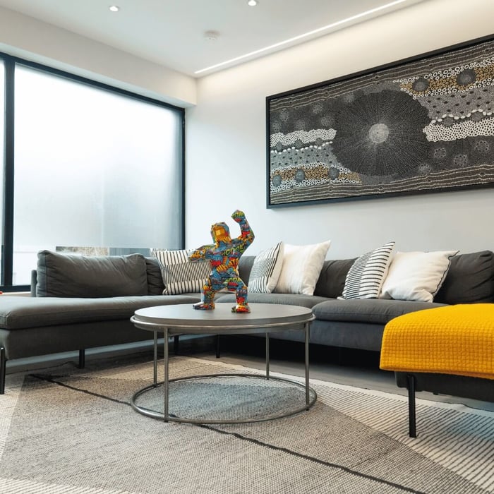 Modern living room featuring a vibrant, colourful geometric pop art gorilla sculpture on a round coffee table, surrounded by a grey sectional sofa, monochrome cushions, and a yellow throw, with a textured abstract wall art in the background.