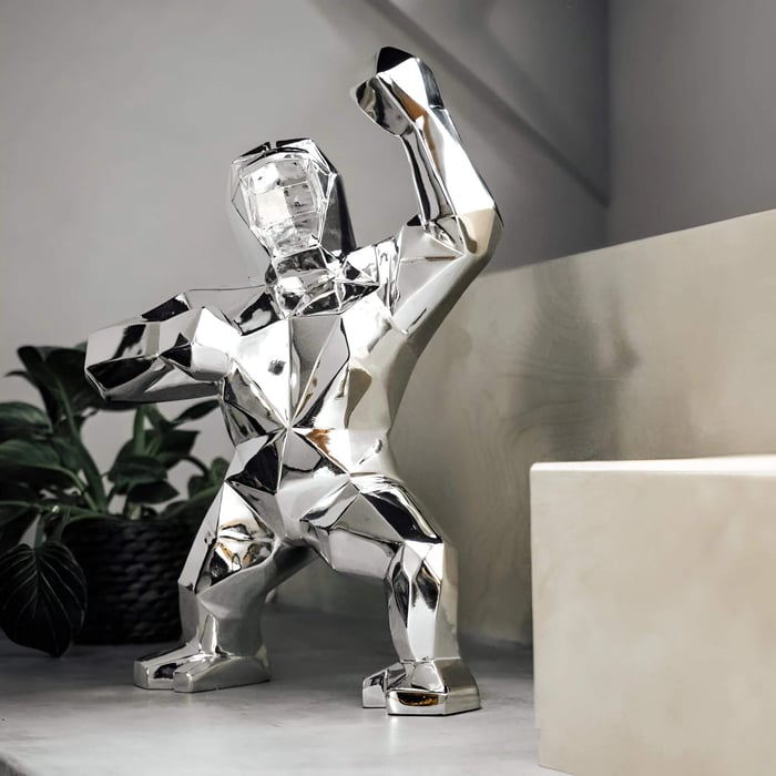 Polished silver geometric gorilla sculpture with a raised fist displayed on a modern shelf, with a potted plant and neutral walls.