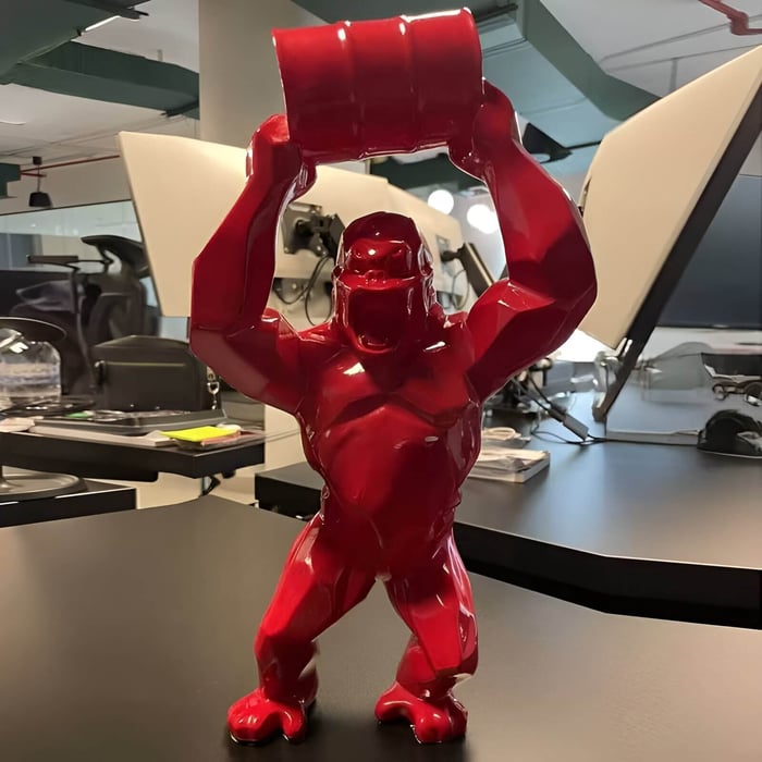 Glossy red geometric gorilla sculpture holding a barrel overhead on a black desk in a modern office.