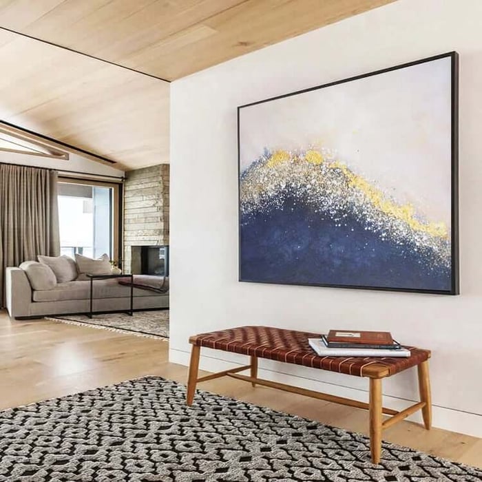 Square abstract painting with a navy-and-gold, wave-like texture on a pale background, hanging above a woven leather bench in a modern living room.
