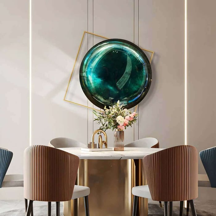 Modern dining area with a round marble and gold table, fluted upholstered chairs, and a large circular teal wall sculpture framed by a tilted gold square above a floral centerpiece.
