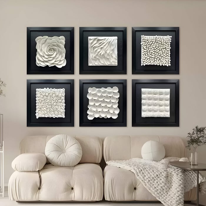 A beige sofa with round cushions is placed beneath a display of six Harmonia 3D Petal Wall Sculptures by Giant Sculptures, each in black frames. These art pieces feature textured white floral and geometric designs. A basket and plant flank the sofa on either side.