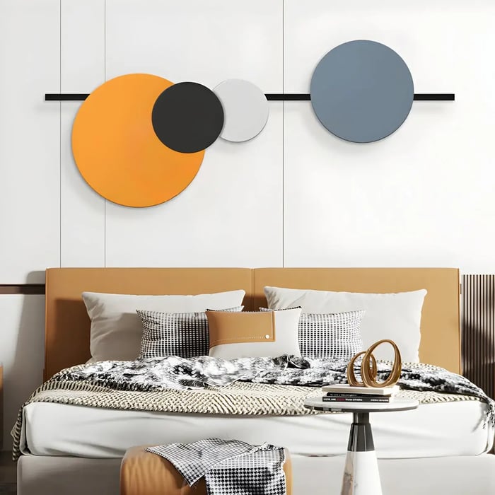 A stylish bedroom with a geometric wall art installation of overlapping circular panels in orange, grey, and black, positioned above a tan upholstered bed.