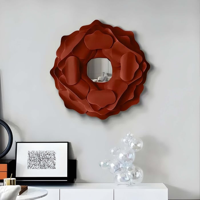A contemporary room showcases the Giant Sculptures Sculptura Mirror, a terracotta red floral round metal wall art. Below, a white console holds framed art and a clear glass bubble sculpture. A white table is partially visible in the foreground.