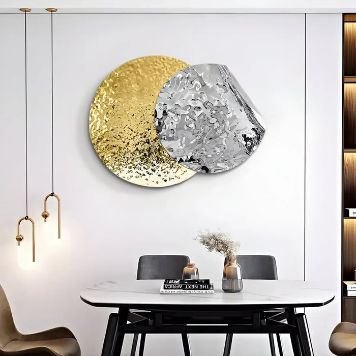 A round metal artwork featuring a gold and silver hammered texture, slightly curled, mounted on a white wall in a dining room with pendant lighting.