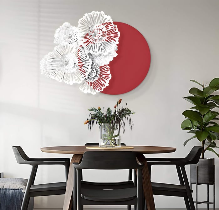 A modern dining room features a round table with four chairs and a flower vase. On the wall, the Fleur Halo Red Round White Metal Floral Decorative Wall Art by Giant Sculptures showcases white metal flowers with a red circle. A tall green plant enhances the serene ambiance.