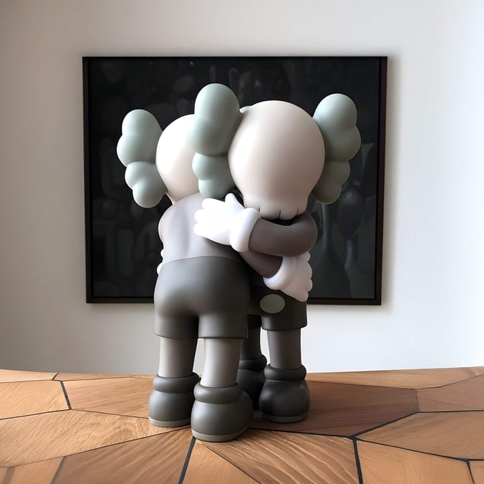 Two stylized resin sculptures with grey tones embrace in the Giant Sculptures Hazelnut Brown Iconify Embrace Figure - 28cm. They stand on a wooden surface against dark-framed art, with exaggerated hands and feet that charm this softly lit home décor piece.