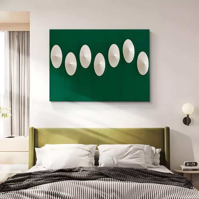 Rectangular green 3D wall art with a sequence of white sculptural discs in rhythmic alignment, mounted above a bed with an olive green headboard in a bright modern bedroom.