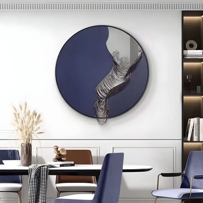 Round navy blue mirror wall art with a sculptural wave-like silver form cascading across the surface, displayed above a modern dining table with mixed-colour chairs and minimalist decor.