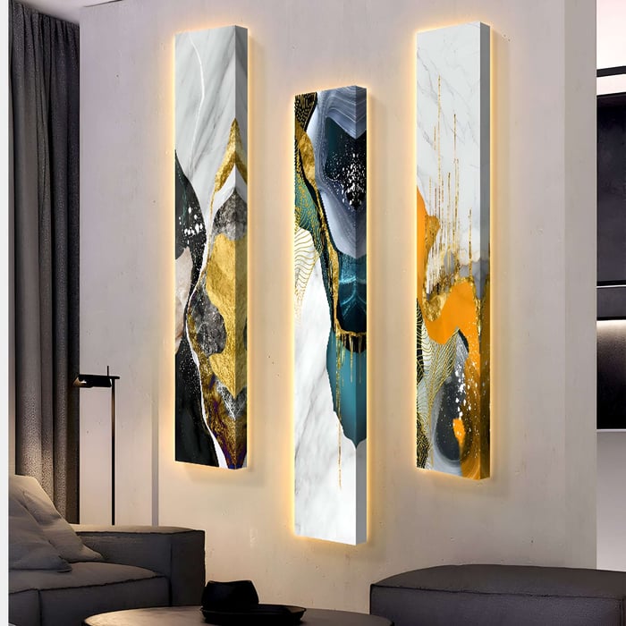Three Floren Amber Glow Canvas LED artworks by Giant Sculptures are displayed on a wall, showcasing gold, white, teal, and orange hues in flowing patterns. A modern sofa is partially visible nearby in the room.