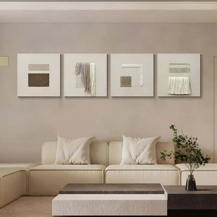 Four square neutral wall artworks in a row above a cream sofa, featuring subtle texture and fibre details.