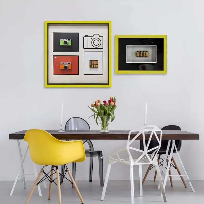 Modern dining room with a wooden table, mixed contemporary chairs, fresh tulips, and two framed 3D wall art pieces featuring bright cameras and film rolls in yellow-accented frames.