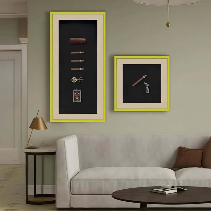 Two lime-framed shadowbox wall artworks displaying cigars and cigar accessories above a cream sofa in a minimalist living room.