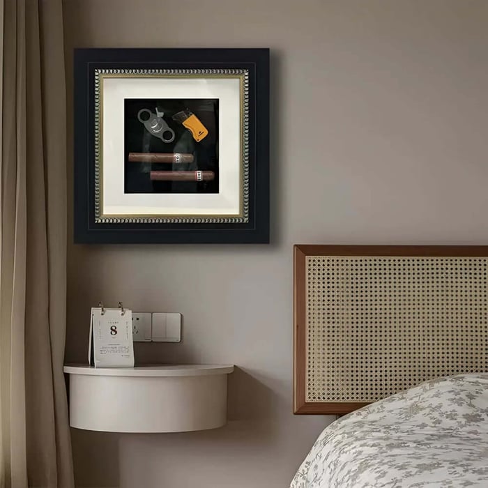 Framed shadowbox wall art with two cigars, a cigar cutter and a lighter above a bedside table.