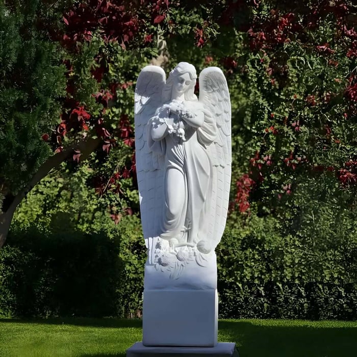 Marble angel sculpture standing on a base and holding a garland of flowers in a green garden.