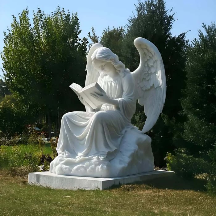 Large marble angel sculpture seated on a cloud reading a book in a garden setting.