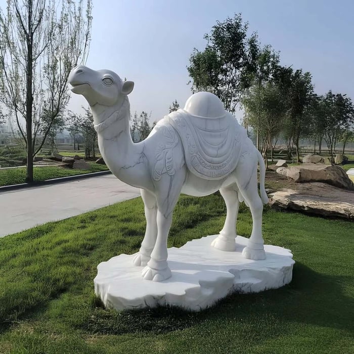 White marble camel statue with carved saddle details, standing on a stone base in a landscaped outdoor area.