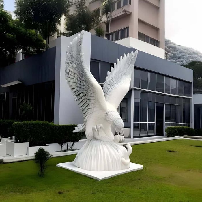 Large white marble eagle sculpture with wings spread, displayed on a lawn outside a modern building with glass windows.