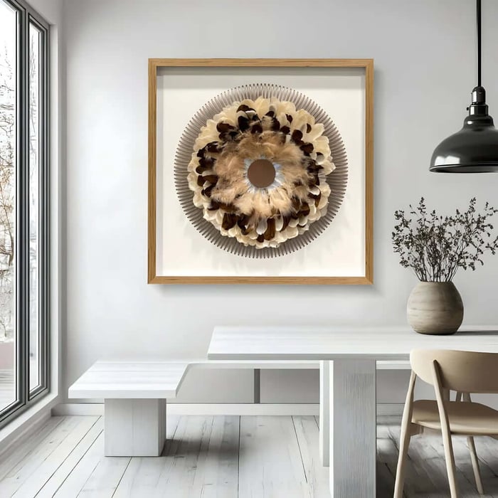 Framed circular feather wall art in neutral tones hanging above a white dining table in a bright, minimalist room.