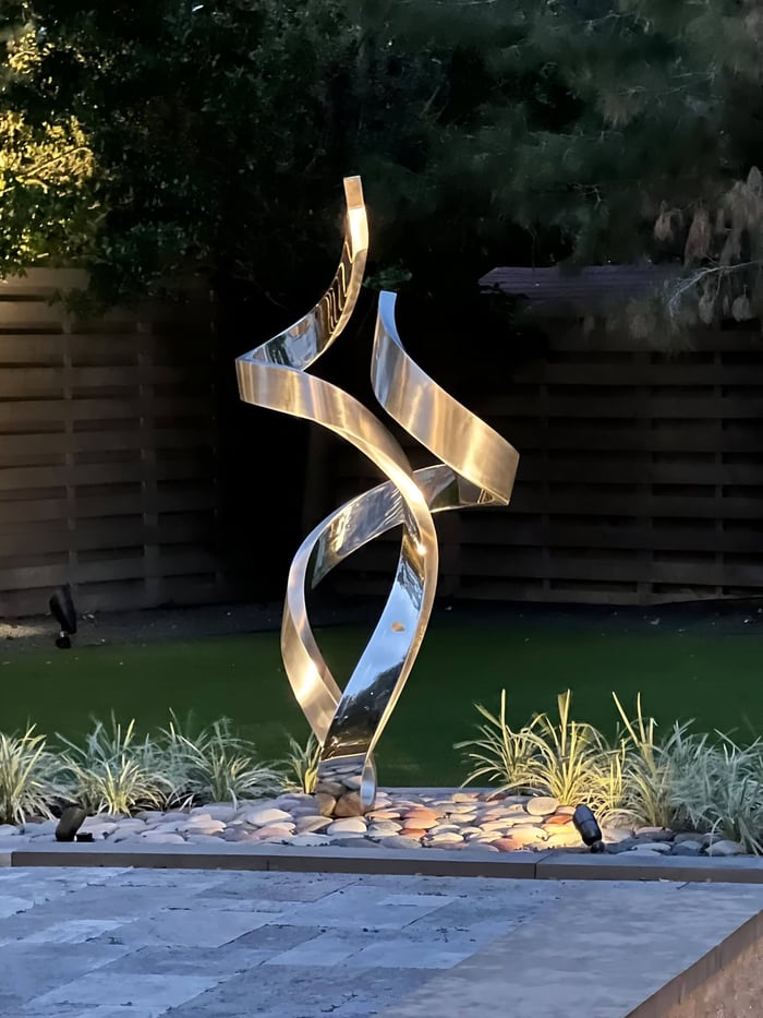 Tall polished metal ribbon sculpture with twisting curves, dramatically lit from below on a bed of river rocks in a landscaped backyard at dusk.