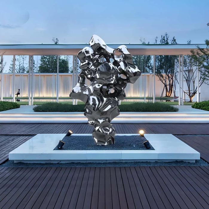 Abstract polished metal sculpture with irregular, organic shapes displayed on a modern outdoor platform in front of a contemporary building.