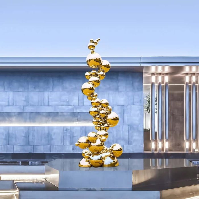 Tall outdoor sculpture made of stacked reflective gold spheres twisting upward on a polished platform in front of a modern gray metal building.