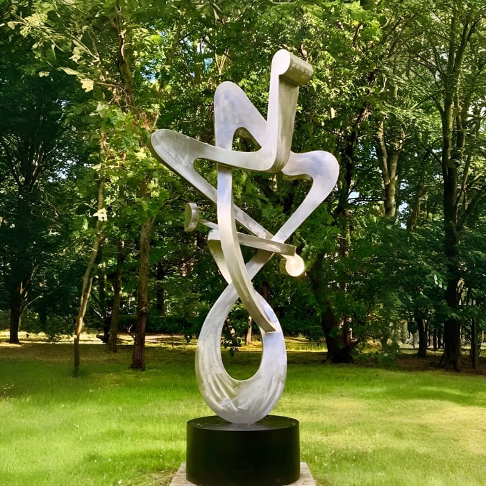 Tall abstract silver metal sculpture with looping, ribbon-like curves mounted on a black cylindrical base in a sunny green park surrounded by trees.