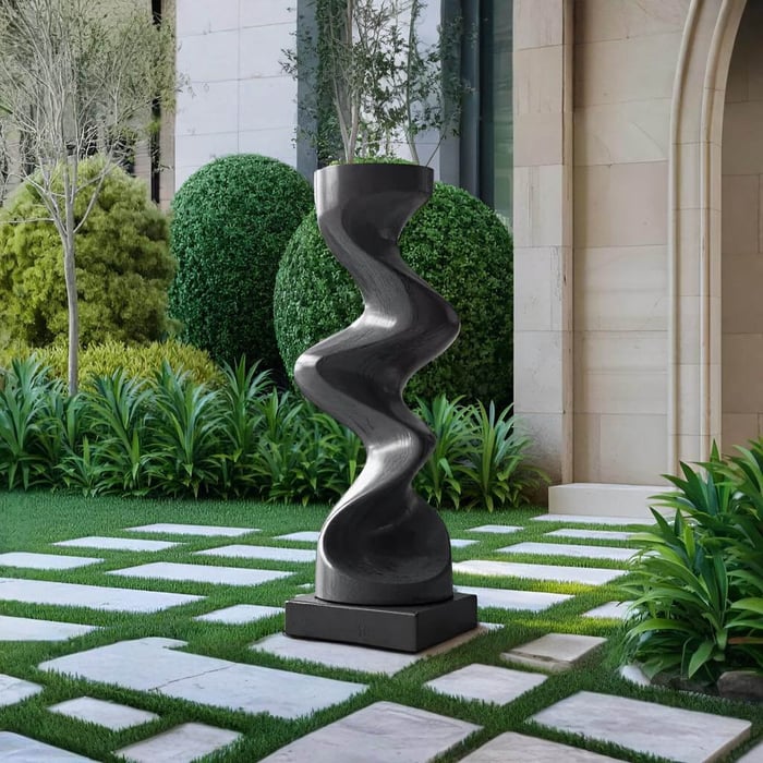 Tall black abstract sculpture with a smooth, twisting hourglass shape, standing on a square base in a manicured garden with stepping stones, lush green plants, and neatly trimmed shrubs.