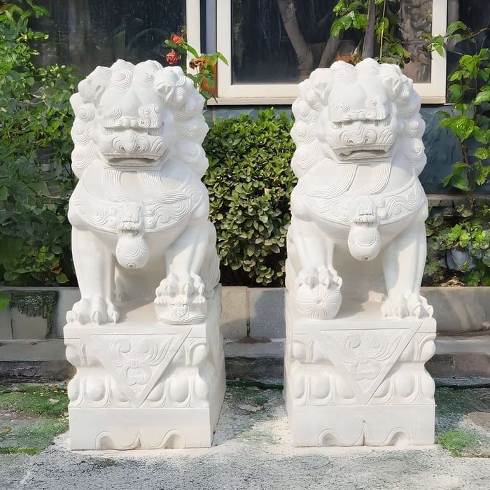 Two Chinese Guardian Lion VI Marble Outdoor Garden Sculptures by Giant Sculptures rest atop square pedestals outside a building. These intricately carved pieces feature detailed elements and decorative patterns, set against a backdrop of greenery and a window.