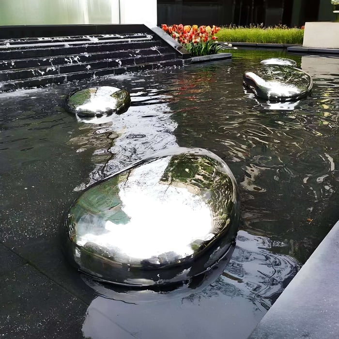 Reflective pebble stainless steel sculptures placed in a water feature, complementing a modern garden.