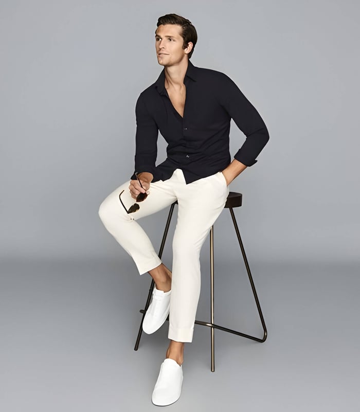 a man wearing white trousers and a black shirt, perched on a stood
