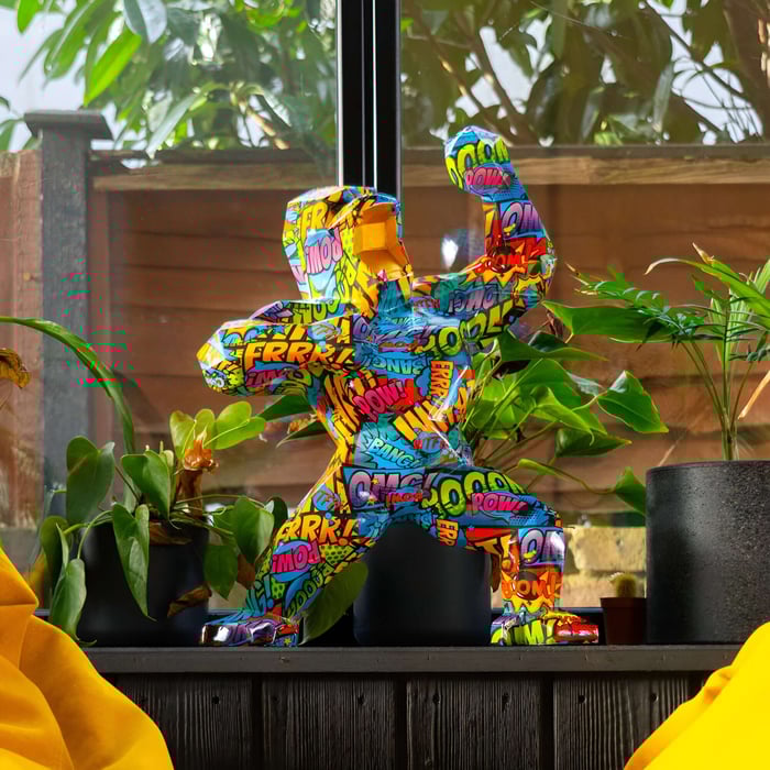 Pop Art gorilla sculpture displayed on a wooden surface surrounded by lush green plants, set against a sunny garden backdrop.