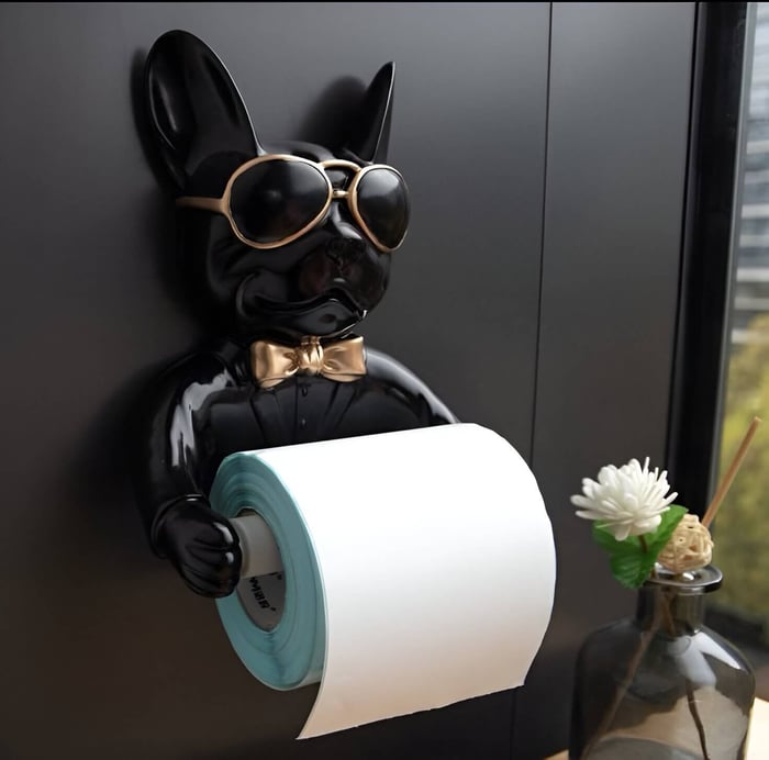 Black resin French bulldog toilet paper holder with gold sunglasses and bow tie, mounted on a bathroom wall, next to a decorative flower vase.