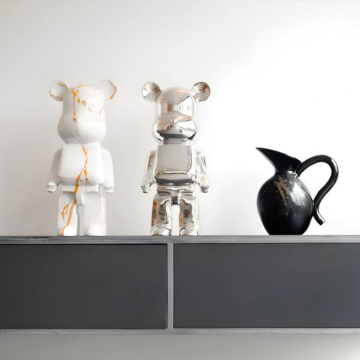 Two bear-shaped sculptures a white one with gold marbling and a reflective chrome one—stand on a minimalist shelf beside a black ceramic pitcher against a white wall.
