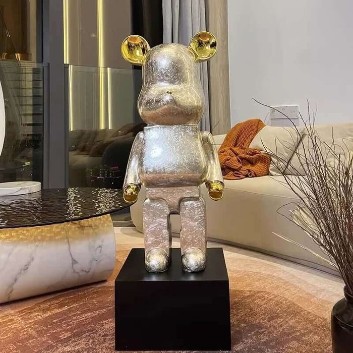 A shiny silver bear-shaped sculpture with gold ears and hands stands on a black pedestal in a modern living room beside a marble coffee table and a beige sofa with an orange throw.