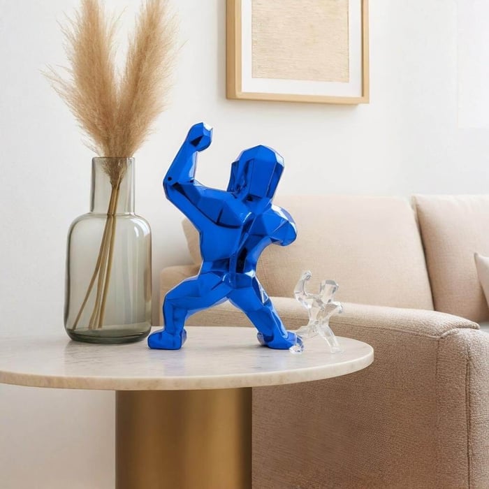 A shiny, faceted blue bear-like sculpture poses on a round marble side table in a bright, neutral living room, next to a tall glass vase with pampas grass and a small clear figurine, with a beige sofa and framed wall art behind it.