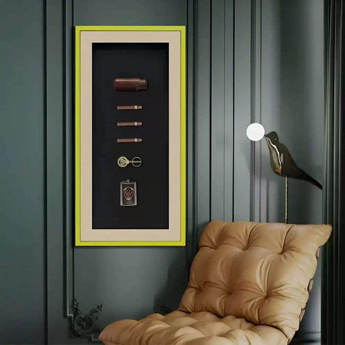 Vertical framed shadowbox with cigar case, cigars and accessories on a dark panelled wall beside a lounge chair.