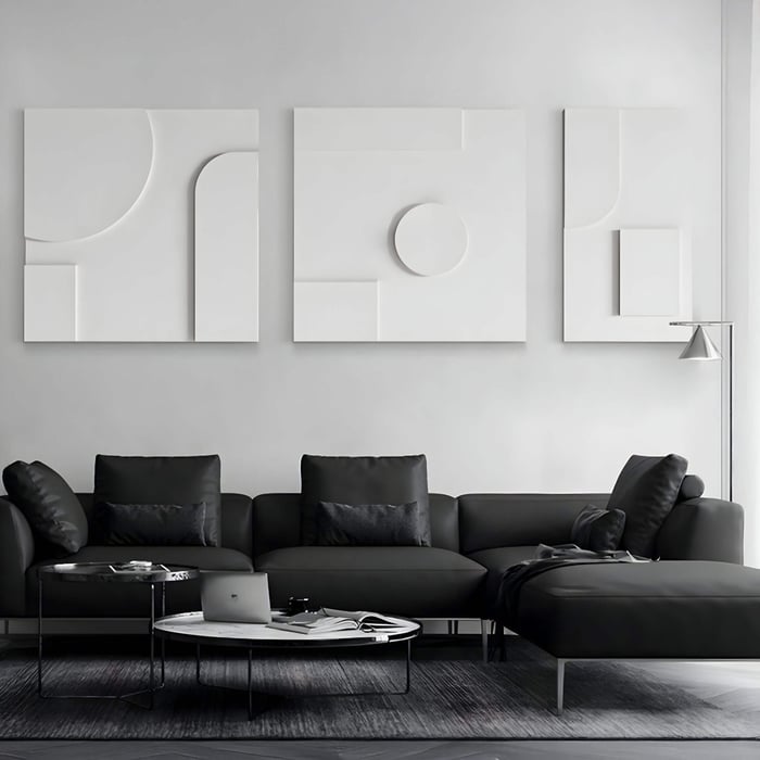Minimalist triptych sandstone wall art in white relief, featuring abstract geometric shapes and clean 3D textures, displayed above a modern black sectional sofa in a monochrome living room.