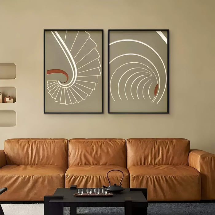 Two framed abstract spiral artworks in beige and white tones hang above a brown leather sofa in a modern living room.