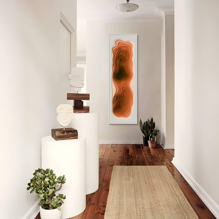 Vertical abstract 3D wall art featuring layered orange and brown carved gradient forms framed in white, displayed in a modern hallway with plants and sculptural accents.